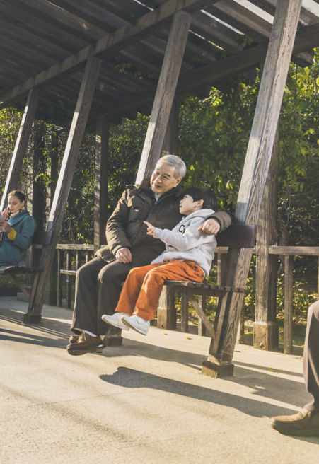 A young boy and elderly man sit on a bench among green trees in China.